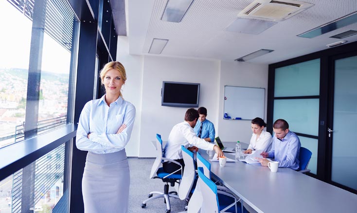 Businesswoman standing in office with a team meeting in the background.